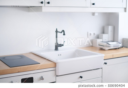 The interior of a kitchen in white tones with a sink, sandwich maker, stove, oven, dining table and a light window 88690404