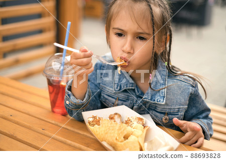 A child bites hard food while sitting at a table in a city cafe outdoors. Lunch in the fresh air in the restaurant patio. Hungry girl with afro pigtails in a denim jacket A child bites hard food while sitting at a table in a city cafe outdoors. Lunch in the fresh air in the restaurant patio. Hungry girl with afro pigtails in a denim jacket 88693828