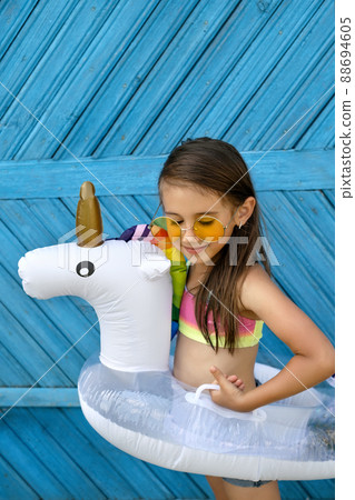 A cute beautiful girl in yellow sunglasses and a swimsuit is standing against a blue wooden plank wall with a circle in the shape of a unicorn. Children's swimming circle for swimming pool A cute beautiful girl in yellow sunglasses and a swimsuit is standing against a blue wooden plank wall with a circle in the shape of a unicorn. Children's swimming circle for swimming pool 88694605