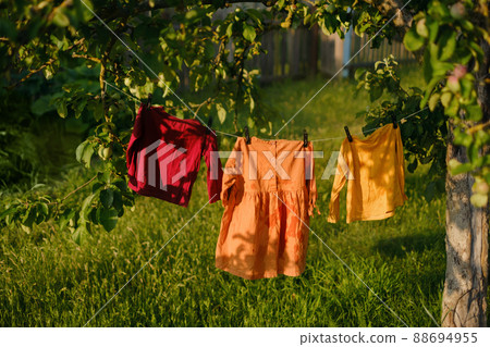 Children's colored clothes hang on a clothesline and dry after washing in nature under a tree in the garden. Clothes flutter in the wind. Hypoallergenic washing powder for babys clothes. 88694955