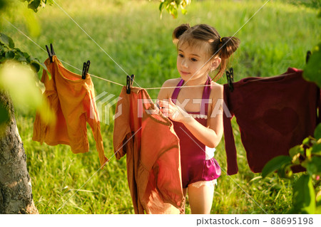 A cute little girl hangs a laundered dress to dry on a clothesline in the garden under a tree. Washing colored clothes with children's hypoallergenic safe washing powder. Rinses for baby clothes 88695198