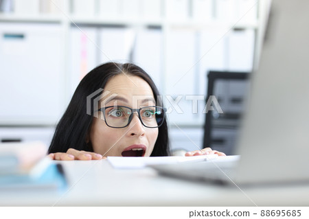 Young businesswoman peering over her desk in office in wide eyed horror or amazement Young businesswoman peering over her desk in office in wide eyed horror or amazement 88695685