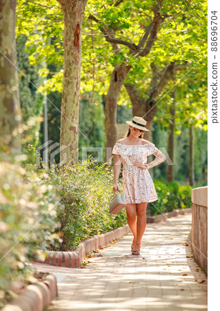 The bright beautiful girl in a light dress and hat walks along alley of trees in Monaco in sunny weather in the summer, handbag in hand The bright beautiful girl in a light dress and hat walks along alley of trees in Monaco in sunny weather in the summer, handbag in hand 88696704