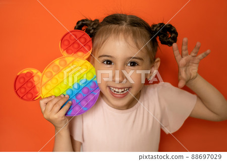 An emotional portrait of a cute little girl on an orange background with a funny hairstyle made of pigtails and a popit toy in her hands. Hand gestures. Pop it toy 88697029