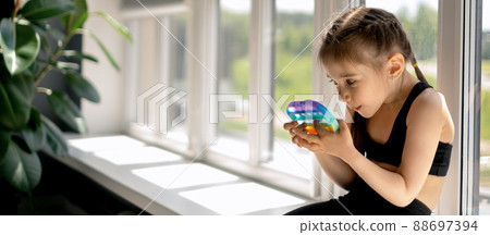 The girl is sitting on the windowsill by the big house and playing with toy with a surprised face. Modern touch toy for the development of fine motor skills of the hands. banner. The girl is sitting on the windowsill by the big house and playing with toy with a surprised face. Modern touch toy for the development of fine motor skills of the hands. banner. 88697394