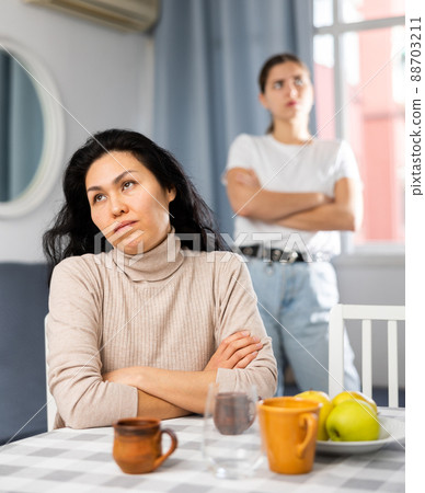 Upset woman sitting at home table with disgruntled woman behind Upset woman sitting at home table with disgruntled woman behind 88703211