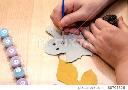 A grown woman draws on fallen leaves. The woman's hands with a paint brush draw a sketchy face on a poplar leaf. Hobbies, creativity, creativity. The bottom of the room. Selective focus. Series part A grown woman draws on fallen leaves. The woman's hands with a paint brush draw a sketchy face on a poplar leaf. Hobbies, creativity, creativity. The bottom of the room. Selective focus. Series part 88705826