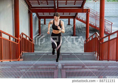 Straight view of young athletic slim beautiful woman running up stairs doing cardio interval training in black fashionable sportwear in the street on summer. Outdoor, sport and healthy concept Straight view of young athletic slim beautiful woman running up stairs doing cardio interval training in black fashionable sportwear in the street on summer. Outdoor, sport and healthy concept 88711131