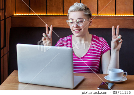 Portrait of beautiful emotional positive girl with blonde short hair in pink t-shirt is sitting in cafe and working on laptop with smile and showing victory sign, looking at laptop. indoor, lifestyle 88711136