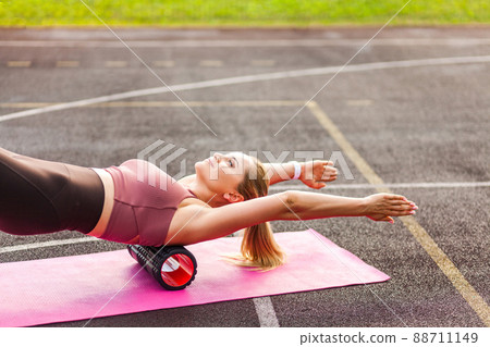 Happy sporty woman training on mat outdoor summer day, using foam roller massager on shoulder and back for relaxation, stretching spine muscles, doing fascia exercise. Health care, workouts routine 88711149