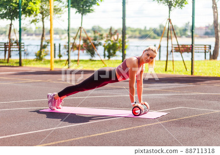 Fit sporty woman training on mat outdoor summer day, performing perfect plank with foam roller massager, doing pilates fitness workouts for muscle build, abs exercises. Health care, sports activity 88711318