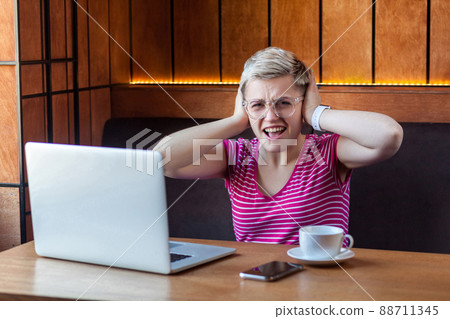 Portrait of attractive young girl freelancer in pink t-shirt is sitting in cafe and working on laptop, covering ears with fingers that not hear to companion. looking at camera Portrait of attractive young girl freelancer in pink t-shirt is sitting in cafe and working on laptop, covering ears with fingers that not hear to companion. looking at camera 88711345