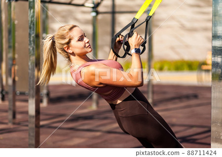 Closeup strong athlete blond woman training outdoor, pulling trx suspension straps, doing upper body workouts for muscle build, fat burning, cardio improvement. Health care and sports activity concept 88711424