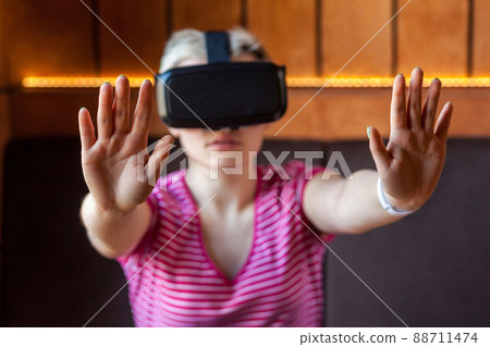 Portrait of serious young adult woman in pink t-shirt sitting in cafe, wearing virtual reality headset and watching video, playing game on simulator raised arms showing stop sign, looking at camera, Portrait of serious young adult woman in pink t-shirt sitting in cafe, wearing virtual reality headset and watching video, playing game on simulator raised arms showing stop sign, looking at camera, 88711474