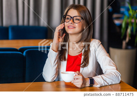 Portrait of happy beautiful stylish brunette young woman in glasses sitting talking on mobile smart phone and toothy smile enjoying. indoor studio shot, cafe, office background. 88711475