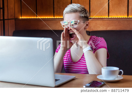 Portrait of angry upset young adult woman with blonde short hair, in pink t-shirt and eyeglasses is sitting in cafe and screaming loud voice to laptop with aggressive face. Indoor, lifestyle Portrait of angry upset young adult woman with blonde short hair, in pink t-shirt and eyeglasses is sitting in cafe and screaming loud voice to laptop with aggressive face. Indoor, lifestyle 88711479