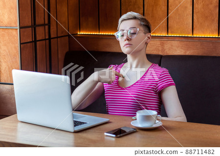 Portrait of confident egoist young girl with short hair in pink t-shirt and eyeglasses is sitting alone in cafe and pointing finger to herself with proud, looking at laptop. Indoor, lifestyle Portrait of confident egoist young girl with short hair in pink t-shirt and eyeglasses is sitting alone in cafe and pointing finger to herself with proud, looking at laptop. Indoor, lifestyle 88711482