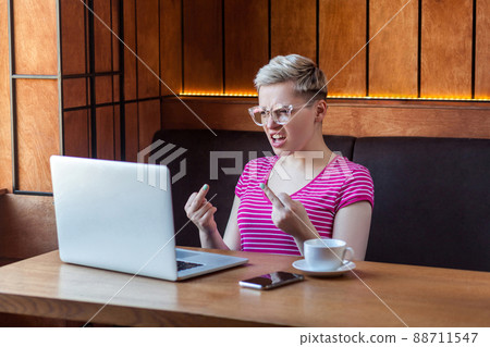 Aggressive unhappy young girl freelancer with blonde short hair, in pink t-shirt and eyeglasses is sitting in cafe and having bad mood are showing fuck sing with finger through a webcam. indoor 88711547