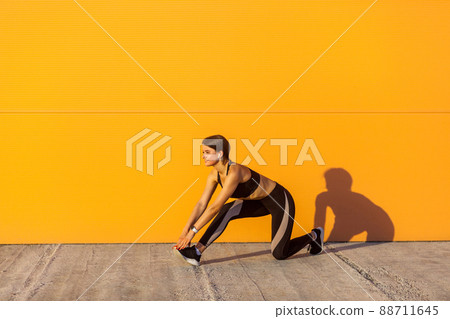 Side view of young beautiful sporty yogi female in black sportwear stretching on the street floor standing on knee, looking straight, Outdoor, orange wall background, sport and healthy concept Side view of young beautiful sporty yogi female in black sportwear stretching on the street floor standing on knee, looking straight, Outdoor, orange wall background, sport and healthy concept 88711645
