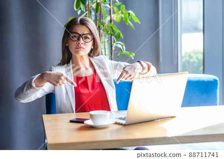 Here and right know. Portrait of serious beautiful stylish brunette young woman in glasses sitting, pointing down and want to come to her quickly. indoor studio shot, cafe, office background. Here and right know. Portrait of serious beautiful stylish brunette young woman in glasses sitting, pointing down and want to come to her quickly. indoor studio shot, cafe, office background. 88711741