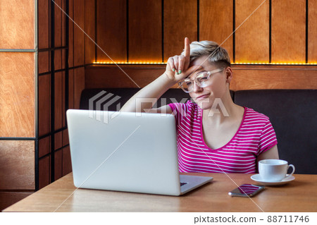 Unhappy rude young girl freelancer with blonde short hair in pink t-shirt and eyeglasses is sitting in cafe and having bad mood are showing hater sing with finger at laptop. indoor, lifestyle Unhappy rude young girl freelancer with blonde short hair in pink t-shirt and eyeglasses is sitting in cafe and having bad mood are showing hater sing with finger at laptop. indoor, lifestyle 88711746