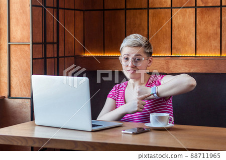 Portrait of upset young bossy girl with blonde short hair in pink shirt and eyeglasses is sitting in cafe and showing checking time, pointing finger to wrist watch, looking at camera indoor Portrait of upset young bossy girl with blonde short hair in pink shirt and eyeglasses is sitting in cafe and showing checking time, pointing finger to wrist watch, looking at camera indoor 88711965