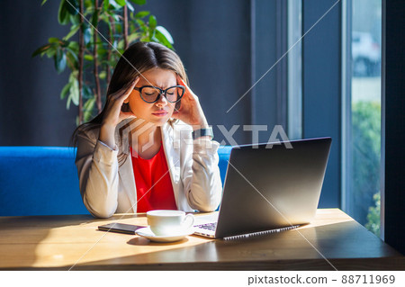 Headache, migraine, confusion, problem or thinking. Portrait of sick brunette young woman in glasses sitting, holding her painful head down and feeling bad. indoor studio shot, cafe, office background Headache, migraine, confusion, problem or thinking. Portrait of sick brunette young woman in glasses sitting, holding her painful head down and feeling bad. indoor studio shot, cafe, office background 88711969
