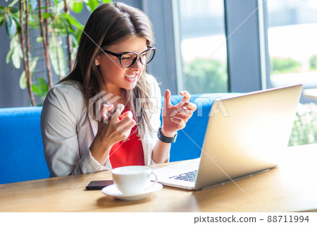 Portrait of angry beautiful stylish brunette young woman in glasses sitting, looking at laptop screen with aggressive face and clenching teeth. indoor studio shot, cafe, office background. Portrait of angry beautiful stylish brunette young woman in glasses sitting, looking at laptop screen with aggressive face and clenching teeth. indoor studio shot, cafe, office background. 88711994
