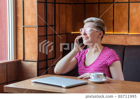 Portrait of beautiful satisfied happy young bussineswoman with blonde short hair in pink t-shirt is sitting in cafe, talking on phone with toothy smile, looking at window. Indoor, healthy lifestyle Portrait of beautiful satisfied happy young bussineswoman with blonde short hair in pink t-shirt is sitting in cafe, talking on phone with toothy smile, looking at window. Indoor, healthy lifestyle 88711995