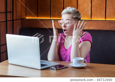 Portrait of unbelievable young girl with short hair in pink t-shirt and eyeglasses is sitting in cafe, working and watching video on laptop, raised hands with shocked face, indoor, lifestyle Portrait of unbelievable young girl with short hair in pink t-shirt and eyeglasses is sitting in cafe, working and watching video on laptop, raised hands with shocked face, indoor, lifestyle 88712000