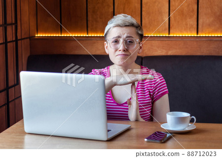 Portrait of warning sad attractive young girl freelancer with blonde short hair is sitting in cafe and looking at camera with raised arms and showing pause gesture, indoor, lifestyle Portrait of warning sad attractive young girl freelancer with blonde short hair is sitting in cafe and looking at camera with raised arms and showing pause gesture, indoor, lifestyle 88712023
