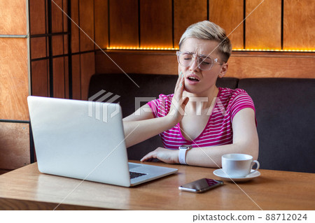 Portrait of unwell young adult woman with short blonde hair in pink t-shirt and eyeglasses sitting in cafe and touching cheek with hand have a high pain, toothache and dentistry. Indoor, health care Portrait of unwell young adult woman with short blonde hair in pink t-shirt and eyeglasses sitting in cafe and touching cheek with hand have a high pain, toothache and dentistry. Indoor, health care 88712024