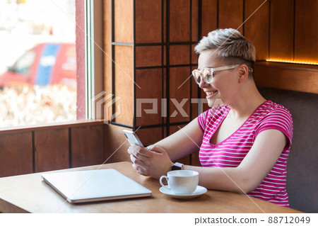 Side view portrait of satisfied happy young blogger with blonde short hair in pink t-shirt is sitting in cafe, holding phone with toothy smile, reading cute message. Indoor, healthy lifestyle 88712049