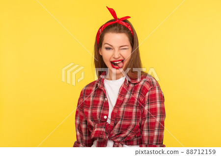 Portrait of optimistic pinup girl in checkered shirt and headband winking to camera with crazy naughty expression, showing tongue out, making faces blinking eye. retro 50's style, studio shot isolated 88712159