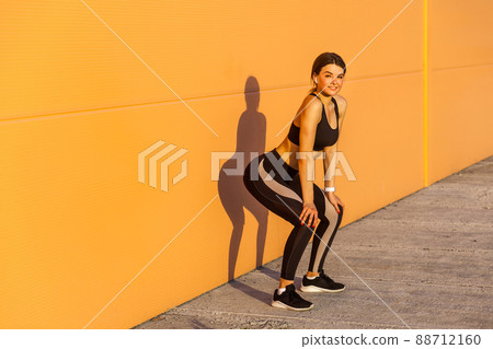Young attractive girl in black sporwear practicing sport exercise on street, standing on squat near orange wall background, take rest after hard workout, outdoor, looking at camera with toothy smile 88712160