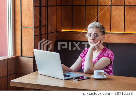 Sadness tired young girl freelancer with short hair, in pink t-shirt is sitting alone in cafe and searching for a work on laptop are in bad mood without good result. looking at camera 88712188