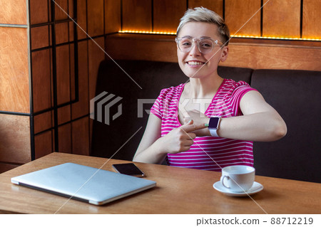 Portrait of beautiful satisfied happy young bussineswoman with blonde short hair in pink t-shirt is sitting in cafe, pointing finger to wrist watch and looking at camera with toothy smile. Indoor, Portrait of beautiful satisfied happy young bussineswoman with blonde short hair in pink t-shirt is sitting in cafe, pointing finger to wrist watch and looking at camera with toothy smile. Indoor, 88712219