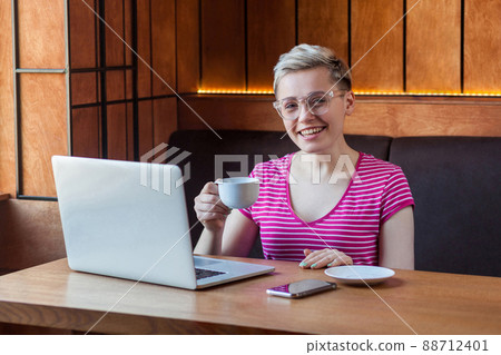 Portrait of beautiful satisfied young bussineswoman with blonde short hair in pink t-shirt is sitting in cafe and drinking coffee and looking at camera with toothy smile. Indoor, healthy lifestyle 88712401