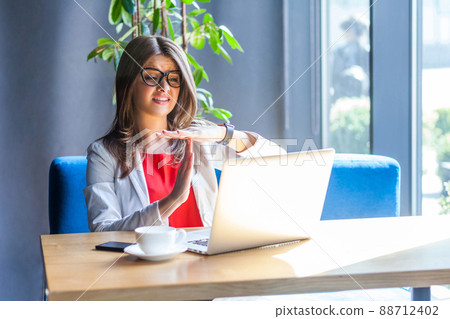 Time out. Portrait of beautiful brunette young woman in glasses sitting, looking at her laptop screen on video call and asking for more time or break. indoor studio shot, cafe, office background. 88712402