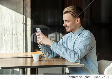 Side view portrait of handsome happy young man in blue denim shirt sitting in cafe and looking and reading message on the phone with toothy smile, looking at the gadget. Indoor, lifestyle 88712427