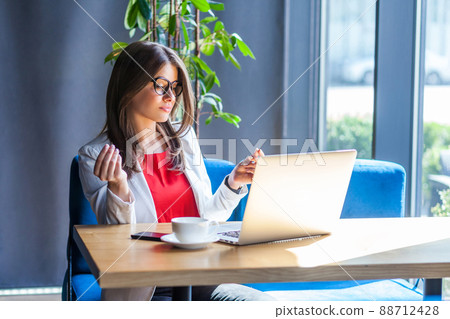 Portrait of beautiful stylish brunette young woman in glasses sitting, looking at her laptop screen on video call and showing money gesture. indoor studio shot, cafe, office background. 88712428