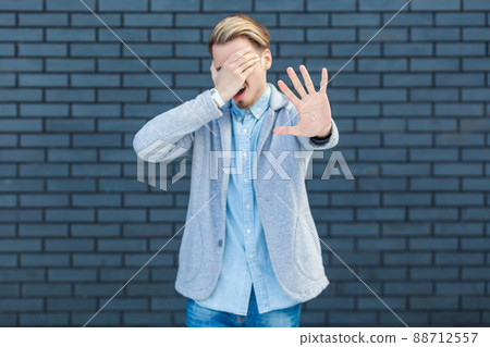 I don't want to look at this. Portrait of ignoring handsome young blonde man in casual style standing, closed his eye and showing stop sign gesture. indoor studio shot on brick wall background. 88712557