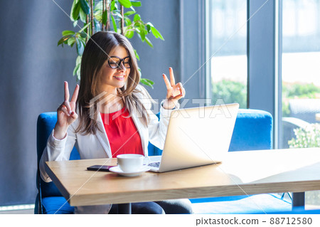 Portrait of happy beautiful stylish brunette young woman in glasses sitting, looking at her laptop monitor on video call with victory peace sign. indoor studio shot, cafe, office background. Portrait of happy beautiful stylish brunette young woman in glasses sitting, looking at her laptop monitor on video call with victory peace sign. indoor studio shot, cafe, office background. 88712580