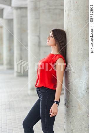 beautiful woman in red blouse and jeans posing while leaning on gray column. outdoor shot. beautiful woman in red blouse and jeans posing while leaning on gray column. outdoor shot. 88712601