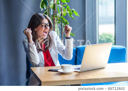 Portrait of happy beautiful stylish brunette young woman in glasses sitting, looking at laptop screen good resualt, screaming and celebrating her victory. indoor studio shot, cafe, office background. Portrait of happy beautiful stylish brunette young woman in glasses sitting, looking at laptop screen good resualt, screaming and celebrating her victory. indoor studio shot, cafe, office background. 88712602