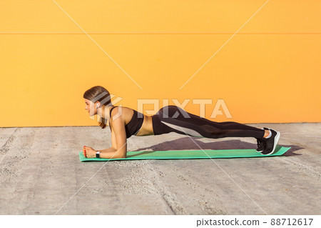 Side view portrait of muscular sporty strong young athletic woman in black sportwear standing on plank position on elbows on green mat. Outdoor orange wall background, sport and healthy concept Side view portrait of muscular sporty strong young athletic woman in black sportwear standing on plank position on elbows on green mat. Outdoor orange wall background, sport and healthy concept 88712617