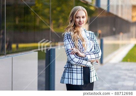 Portrait of attractive confident businesslady with long blond hair in plaid jacket and skirt standing with crossed arms and looking at camera with toothy smile. outdoors, near business center building 88712698