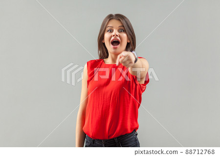 Wow, you are unbelievable. Portrait of surprised beautiful brunette young woman in red shirt standing, pointing and looking at camera with amazed face. indoor, studio shot, isolated on gray background 88712768