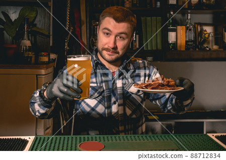 Friendly bearded bartender offering glass of beer with snacks in pub 88712834