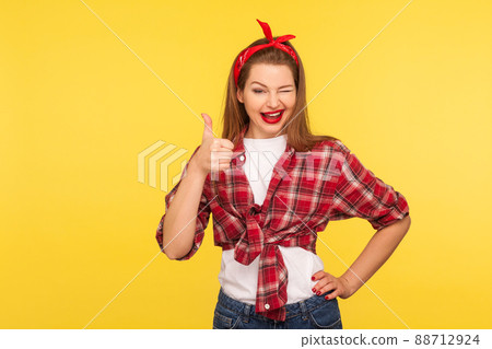 Good job, like it. Portrait of happy coquette pinup girl in checkered shirt and headband winking to camera and showing thumbs up. retro vintage 50's style, studio shot isolated on yellow background 88712924